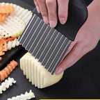 Close-up of the stainless blades of the crinkle cutter slicing a potato into ridged sticks on a slate board.