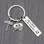 Same camping keychain photographed on a dark grey wood-grain surface, charms and quote bar clearly lit.