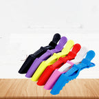Seven plastic hair sectioning clips fanned out in black, purple, yellow, red, pink, white and blue on a pale wood surface.